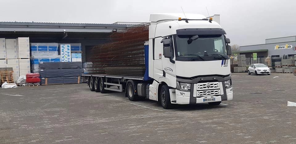 Tracteur Renault T attelé à une semi-remorque plateau chargée d’armatures métalliques, devant un entrepôt